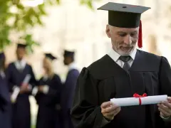 Starszy pan w todze z dyplomem zastanawia się, czy studia liczą się do stażu pracy. W tle inni absolwenci.