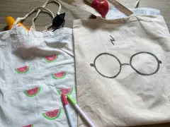 Zwei jute beutel bemalen: einer mit Wassermelonen, der andere mit Harry Potters Brille. Sonnencreme, Sonnenbrille und Stifte liegen daneben.