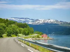 En slingrande väg längs en fjord i Norge, med snöklädda berg i bakgrunden. En röd stuga vid vattnet. Perfekt för en roadtrip norge rutt.