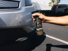 A hand sprays bug remover onto a car bumper, tackling stubborn bugs from a road trip.