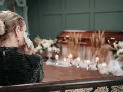 Kobieta ociera łzy chusteczką przy trumnie, otoczonej kwiatami i świecami. W tle widać wyrazy współczucia po niemiecku.