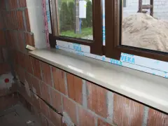 Montaż parapetów zewnętrznych po ociepleniu. Nowy, jasny parapet lśni pod brązową ramą okna, a ceglana ściana czeka na dalsze prace.