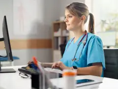 Pielęgniarka w niebieskim uniformie pracuje przy komputerze. Jej specjalizacja pielęgniarska to 5 lat doświadczenia.