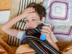 Chory chłopiec leży w łóżku z ręką na czole i trzyma inhalator.