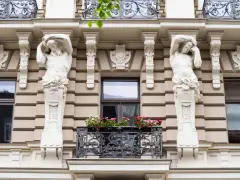 Dwie rzeźby atlantesów podtrzymujące balkon, przykład secesji w sztuce, zdobią fasadę kamienicy.