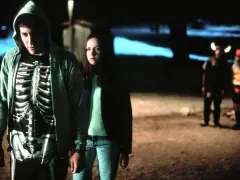 Donnie Darko w bluzie z kapturem i koszulce z nadrukiem szkieletu, obok dziewczyny. W tle dwie postacie w kostiumach.