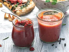 Dwa słoiki domowego sosu na pizzę, jeden z łyżką pełną aromatycznej mieszanki. Obok kawałek pizzy i przyprawy.