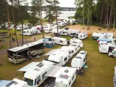 Duży zlot caravaningowy nad jeziorem. Wiele kamperów i przyczep kempingowych zaparkowanych na trawie, otoczonych drzewami.