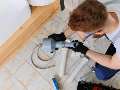 Hydraulik pracuje nad odpowietrzeniem pionu sanitarnego, zgodnie z normą. Widoczny jest zlew i narzędzia.