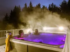 Zwei Frauen stoßen mit Champagner in einem beleuchteten Whirlpool an, umgeben von Dampf und Bäumen. Ein perfektes Wochenende mit Gott.