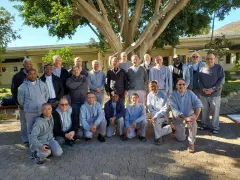 A group of missionaries of charity fathers pose for a photo outdoors, standing and kneeling under a large tree.