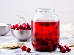 Duży słoik wypełniony rubinowym kompotem z wiśni, obok miska świeżych owoców i cukru.
