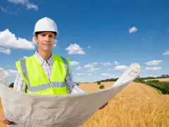 Inżynier w kasku i kamizelce odblaskowej trzyma plany na tle pola. To wizualizacja, jak działka rolno budowlana co to znaczy, czyli połączenie upraw z możliwością zabudowy.
