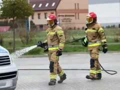Dwóch strażaków w akcji. Jeden z nich polewa wodą samochód, drugi niesie wąż. Ciekawe, ile zarabia strażak za taką pracę.