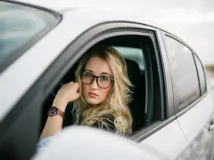 Blondynka w okularach siedzi za kierownicą białego samochodu. Pokazuje, jak nosić okulary, by wyglądać stylowo i pewnie.