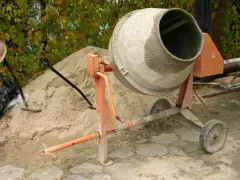 Betoniarka 200l czeka na piasek i cement. Ile cementu na betoniarkę 200l? Obok łopata i kapelusz.