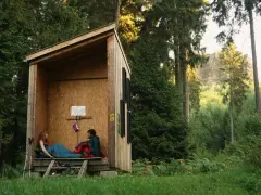 Zwei Personen entspannen sich in einer einfachen Holzhütte, bereit für wild camping. Umgeben von hohen Bäumen und üppigem Grün.