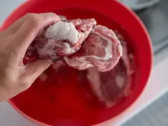 Ręka wkłada kawałki surowego mięsa do czerwonej miski z wodą, pokazując, jak szybko rozmrozić mięso.