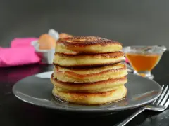 Pila di pancake senza latte, dorati e soffici, con sciroppo d'acero e un tovagliolo rosa sullo sfondo.