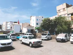 Taxis warten auf Fahrgäste in einer sonnigen Stadt in Marokko. Weiße Gebäude und Bäume säumen die Straße.