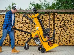Mężczyzna w kasku i okularach ochronnych przygotowuje się do pracy z szybką łuparką do drewna. Za nim piętrzą się stosy drewna opałowego.