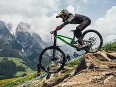 Troy Brosnan in mid-air on a green downhill bike, with a dramatic mountain backdrop.