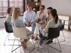 Grupa ludzi siedzi w kręgu, słuchając terapeuty. To scena z ośrodka dla uzależnionych, gdzie wsparcie i zrozumienie pomagają w powrocie do zdrowia.