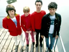 Die Band Talking Heads steht auf einem Holzsteg. David Byrne trägt ein rotes Hemd, Tina Weymouth ein pinkes Outfit.