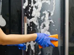 Main bleue, éponge orange pour nettoyer paroi douche. Mousse et bulles sur la paroi vitrée.