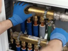 Ręce w niebieskich rękawiczkach dokręcają kluczem mosiężny element instalacji hydraulicznej w domu.