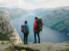 Två vandrare med stora ryggsäckar står på en klippa och blickar ut över en fjord i Norge. Perfekt för camping i Norge.