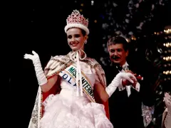 Uśmiechnięta Miss International w koronie i białej sukni, z szarfą "Miss Polonia". Obok niej mężczyzna w smokingu.