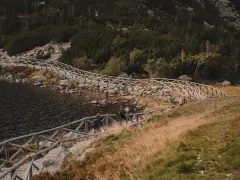 Dwie osoby idą kamiennym szlakiem przy jeziorze, otoczonym drewnianym ogrodzeniem. W oddali widać las i góry.