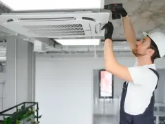 Technik w kasku montuje klimatyzator kasetonowy na suficie.