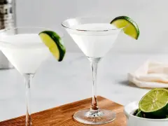 Dwa kieliszki z białym drinkiem, ozdobione plasterkami limonki. Obok miseczka z limonkami i shaker. Idealny kamikaze shot na gorący wieczór.