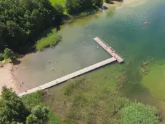 Letnie popołudnie nad jeziorem Hańcza. Ludzie kąpią się i odpoczywają na plaży, inni spacerują po drewnianym pomoście.