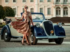 Elegancka kobieta w kwiecistej sukience i okularach przeciwsłonecznych opiera się o zabytkowy samochód. Stylizacja nawiązuje do mody lat 20.