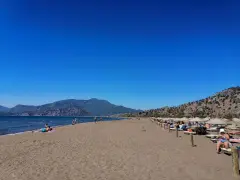Słoneczna plaża w Sarigerme, z turkusowym morzem, górami w tle i rzędami leżaków pod słomianymi parasolami.