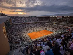 Widok na pełny stadion podczas meczu na kortach ziemnych. Zachodzące słońce oświetla trybuny pełne widzów, tworząc atmosferę emocjonującego **Roland Garros live**.