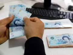Ręce liczą banknoty 10 dinarów, tunezyjska waluta. Na stole leżą stosy pieniędzy, a w tle widać klawiaturę komputera.