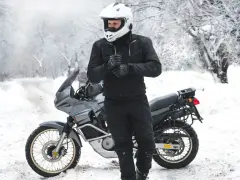 Motociclista con casco bianco e abbigliamento invernale nero, pronto per un'avventura in moto inverno su una strada innevata.