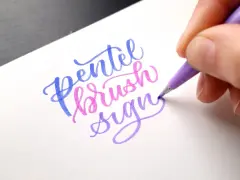 En hand skapar vacker brush lettering med en lila penna. Orden "Pentel brush sign" framträder i blått, rosa och lila.
