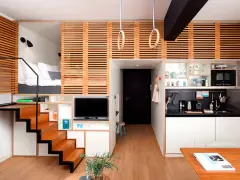 Estudio moderno de piso 25 metros cuadrados con cama elevada, cocina compacta, zona de TV y escaleras de madera.