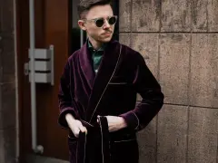 Elegancki mężczyzna w bordowej bonżurce z aksamitu, w okularach przeciwsłonecznych, stoi przy kamiennej ścianie.