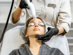 Zabieg laserem na twarz. Fizjoterapeuta wykonuje zabieg, uwzględniając przeciwwskazania. Pacjentka ma okulary ochronne.