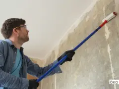 Mann mit Schutzbrille trägt mit langem Stiel und Rolle Öko Tiefengrund auf eine rohe Wand auf.