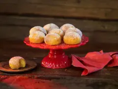 En röd tårtfat med lussebullar med kvarg, pudrade med socker. En ensam bulle ligger på en träplatta.