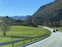 En bil kör på en slingrande väg genom ett grönt landskap med berg och hus. En perfekt start på en semester i Norge.