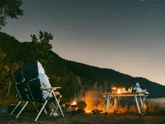 Personne assise près d'un feu de camp sous un ciel étoilé, avec une table de camping et un lac.