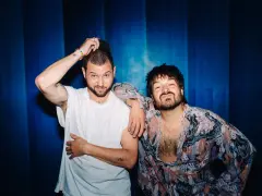 Zwei unabhängige Künstler, einer mit weißem T-Shirt und Kappe, der andere in einem gemusterten Hemd, posieren vor einem blauen Hintergrund.